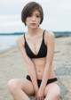 A woman in a black bikini sitting on a rock on the beach.