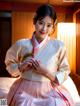 A woman in a pink and white hanbok sitting on a bed.