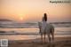 A woman riding a white horse on the beach at sunset.