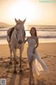 A woman in a white dress standing next to a white horse on the beach.