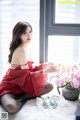 A woman in a red dress sitting on a bed next to a vase of flowers.